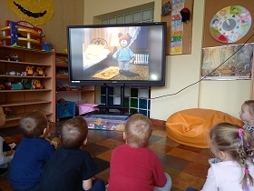 Dzieci siedzą przed ekranem monitora i oglądają bajkę. Na ekranie widać kukiełkę misia.