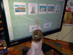 Przed tablicą multimedialną stoi dziewczynka, palcem rysuje linie od zdjęcia Krakowa do napisu Polska.