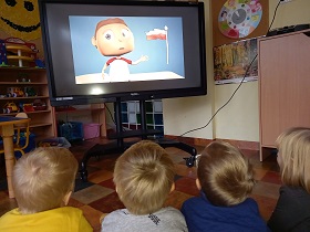 Przed tablicą siedzą dzieci i patrzą w ekran monitora. Na ekranie widać rysunkową postać chłopca, który wskazuje na biało – czerwoną flagę.