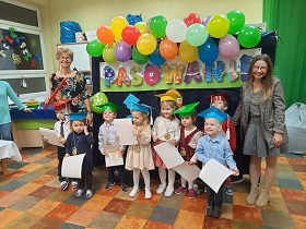 Pani Dyrektor oraz nauczycielka stoją obok dzieci w czerwonych, żółtych, zielonych oraz niebieskich biretach i trzymających dyplomy. Za nimi napis pasowanie i kolorowe balony.