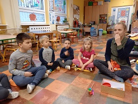 Dzieci siedzą i patrzą w stronę osoby dorosłej, która dłonią pokazuje jak należy ułożyć usta.