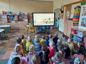 Dzieci siedzą na matach i spoglądają na monitor, na którym wyświetlana jest bajka.