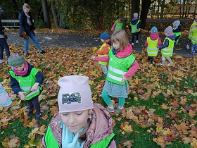 Dzieci w zielonych i żółtych kamizelkach oraz pani w czarnej kurtce, zbierają liście z drzew