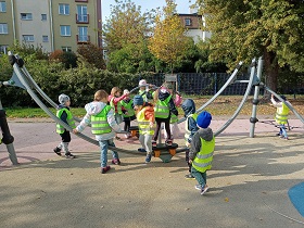 Dzieci w zielonych i żółtych kamizelkach bawią się na placu zabaw na metalowych zabawkach. 