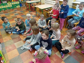 Dzieci siedzą na podłodze i krzesełkach i trzymają w rękach kołatki. Uderzają nimi o dłonie.