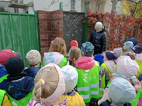 Dzieci w zielonych i żółtych kamizelkach oraz pani w czarnej kurtce stoją przed bramą metalową z dziurami.