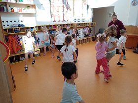 Dzieci w strojach gimnastycznych biegają po sali. Trzymają w rękach czapeczki do ćwiczeń.