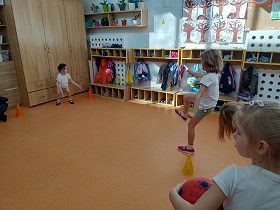 Dziewczynka stoi przy szafkach, między pachołkami. Druga dziewczynka kopie do niej piłką, a trzecia się temu przygląda i trzyma w rękach piłkę.