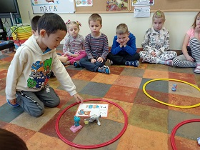Chłopiec na środku koła dzieci, klęczy przy czerwonej obręczy, w której znajdują się dwie zabawki i plansza z cyfrą 2. Na podłodze znajduję się jeszcze żółta i czerwona obręcz. Pozostałe dzieci przyglądają mu się.
