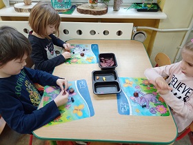 Trójka dzieci siedzi przy stoliku . Na stoliku znajdują się maty, na których dzieci lepią z plasteliny.