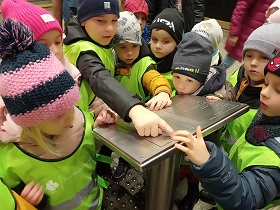Dzieci stoją w grupce i dotykają palcami tablicy dla niewidomych.