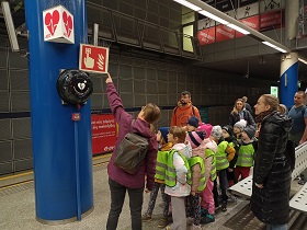 Dzieci wraz z dorosłymi stoją na stacji metra. Pani w fioletowej kurtce pokazuje palcem na tablice ostrzegawcze z ogniem i z serduszkiem.