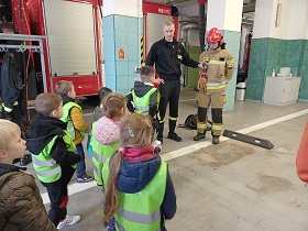 Dzieci w kamizelkach odblaskowych spoglądają na strażaków. Jeden z nich ubrany jest w strój strażaka, który jedzie na akcje ratowniczą i ma na sobie hełm.