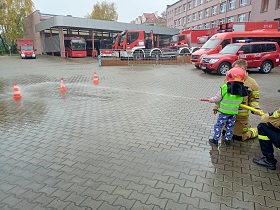 Dziecko w hełmie strażaka celuję wężem strażackim z wodą w pachołek. W tle stoją samochody straży pożarnej. 