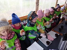 Dzieci w odblaskowych kamizelkach siedzą w jednym rzędzie i jedzą bułki. Przed nimi na stoliku leżą kawałki ręcznika papierowego oraz brązowe kubki.