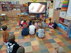 Dzieci siedzą na podłodze przed monitorem, na którym wyświetlane są kredki.