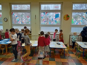 Dzieci chodzą wokół stołów, na których leżą kartki z rysunkami.
