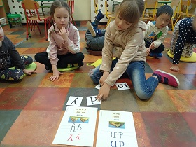 Dziewczynka w beżowej bluzie w białe kropki układa wyraz z rozsypanych na podłodze liter. Przed nią leżą dwie plansze z literą D i Y. Za nią siedzą dzieci na poduszkach i się jej przypatrują.