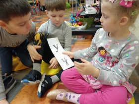 Dwóch chłopców i dziewczynka siedzą na poduszkach i przypatrują się kartce z napisaną literą Y, małą i wielką, którą trzyma w dłoniach dziewczynka.