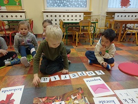 Chłopiec układa słowo koperta z rozsypanych liter. Za nim siedzą dzieci na poduszkach. Przed chłopcem znajdują się plansze z literą k, na których widnieją napisy: kokarda, koperta, korale.