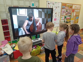 Czwórka dzieci stoi przed monitorem i ogląda pana, który gra na kontrabasie. 