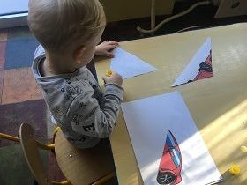 Chłopiec siedzi przy stoliku i przykleja kawałki ilustracji z czerwonym samochodem. 