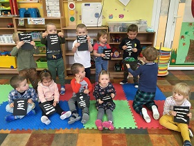 Dzieci siedzą i stoją, trzymając w dłoniach czarne kartki z przyklejonymi plasteliną pasami.