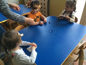 Trójka dzieci siedzi przy stole: po lewej stronie dziewczynka z warkoczykiem i kokardą we włosach w białej bluzce, chłopca w pomarańczowej bluzce, a po prawej stronie dziewczynka w białej bluzce w żółte paski. Dzieci lepią grzyby z plasteliny i kasztanów.