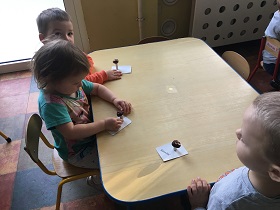 Trójka dzieci siedzi przy stole i lepi grzyby z plasteliny i kasztanów. W środku siedzi dziewczynka w bluzce z Elsą "Z krainy Lodu".