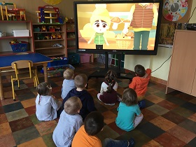 Grupka dzieci siedzi na podłodze i ogląda filmik edukacyjny o grzybach jadalnych i trujących na monitorze.