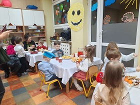 Przy stołach siedzą dzieci z rodzicami i jedzą poczęstunek przygotowany przez rodziców.