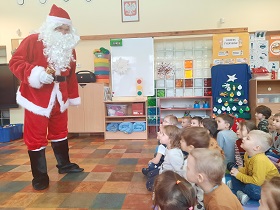 Mikołaj stoi przed dziećmi siedzącymi na podłodze i mówi do nich.