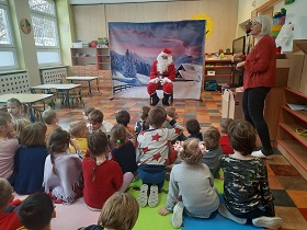 Mikołaj siedzi na krześle, przed nim siedzą dzieci na podłodze i śpiewają piosenkę.