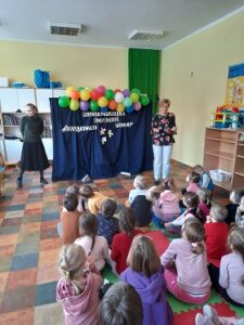 Na środku stoi granatowa dekoracja z kolorowymi balonami, obok stoi Pani Dyrektor oraz nauczycielka, przed dekoracją siedzą dzieci.