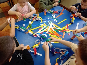 Przy stole siedzą dzieci, na stole położone są kolorowe paski papieru. Dzieci sklejają paski w kółka i tworzą łańcuch.