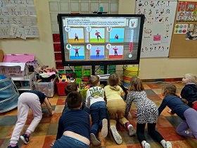 Dzieci klęczą przed monitorem, na którym wyświetlana jest kostka ruchowa z zestawem ćwiczeń.