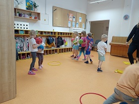 Dzieci biegają po sali z piłkami. Są przebrane w stroje gimnastyczne. Na podłodze leżą obręcze i koszulki.