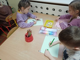 Troje dzieci siedzi przy stoliku i rysuje kredkami i flamastrami coś na kartkach.