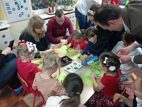 Dzieci wraz z rodzicami siedzą przy stolikach i malują farbami po zielonych kartkach. Jedna pani dolewa farby na paletę.