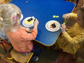 Dwie dziewczynki wykonują prace plastyczną przy stoliku. Jedna ma na sobie bluzkę w kolorze pomarańczowym, a druga w żółtym.