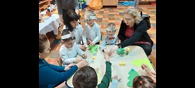Przy stoliku siedzą dzieci i rodzice. Przed każdym dzieckiem leży choinka na którą dzieci naklejają kolorowe elementy.