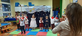 Siedmioro dzieci ubranych świątecznie, tańczą do piosenki. W tle widać świąteczną dekoracje.