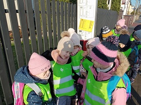 Dzieci stoją na przystanku autobusowym. Mają na sobie kurtki i czapki oraz odblaskowe, zielone kamizelki. Na plecach mają plecaki. 