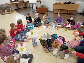 Dzieci siedzą na podłodze w sali i zjadają bułki. Przed nimi stoją kolorowe kubki z herbatą.