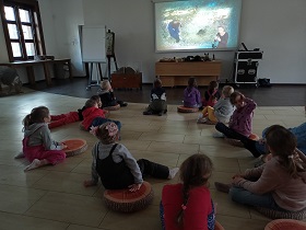 Dzieci siedzą na podłodze w sali i oglądają film, wyświetlany na projektorze.