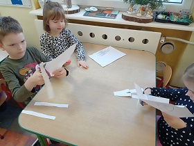 Trójka dzieci siedzi przy stoliku i wycina paski nożyczkami.