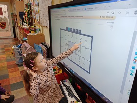 Dziewczynka stoi przy monitorze i dotyka go palcem. Na monitorze widnieją puzzle. 