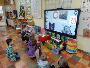 Dzieci siedzą przed monitorem. Na monitorze widnieje napis: W krainie zegarów. Przedstawione są też fotografie różnych rodzajów zegarów. 
