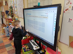 Chłopiec stoi przy monitorze i wskazuje na niego palcem. Na monitorze widać memory zegarowe. 