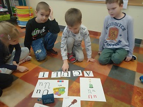 Chłopiec układa słowo z rozsypanki literowej. Przed nim znajdują się planszę z literką U, głośnik oraz obrazek gitary. Inne dzieci przyglądają się temu co robi chłopiec.